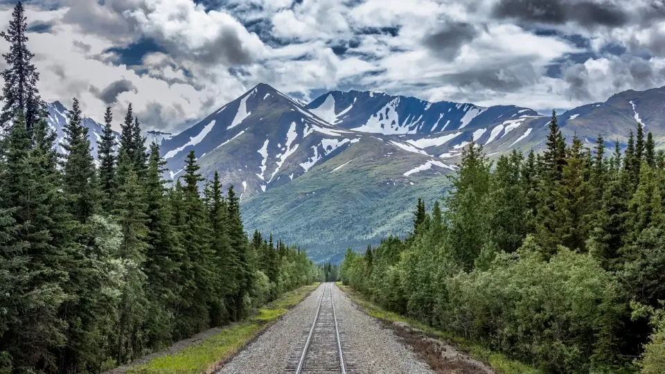 On a railroad track with evergreen trees and snowcapped mountains ahead